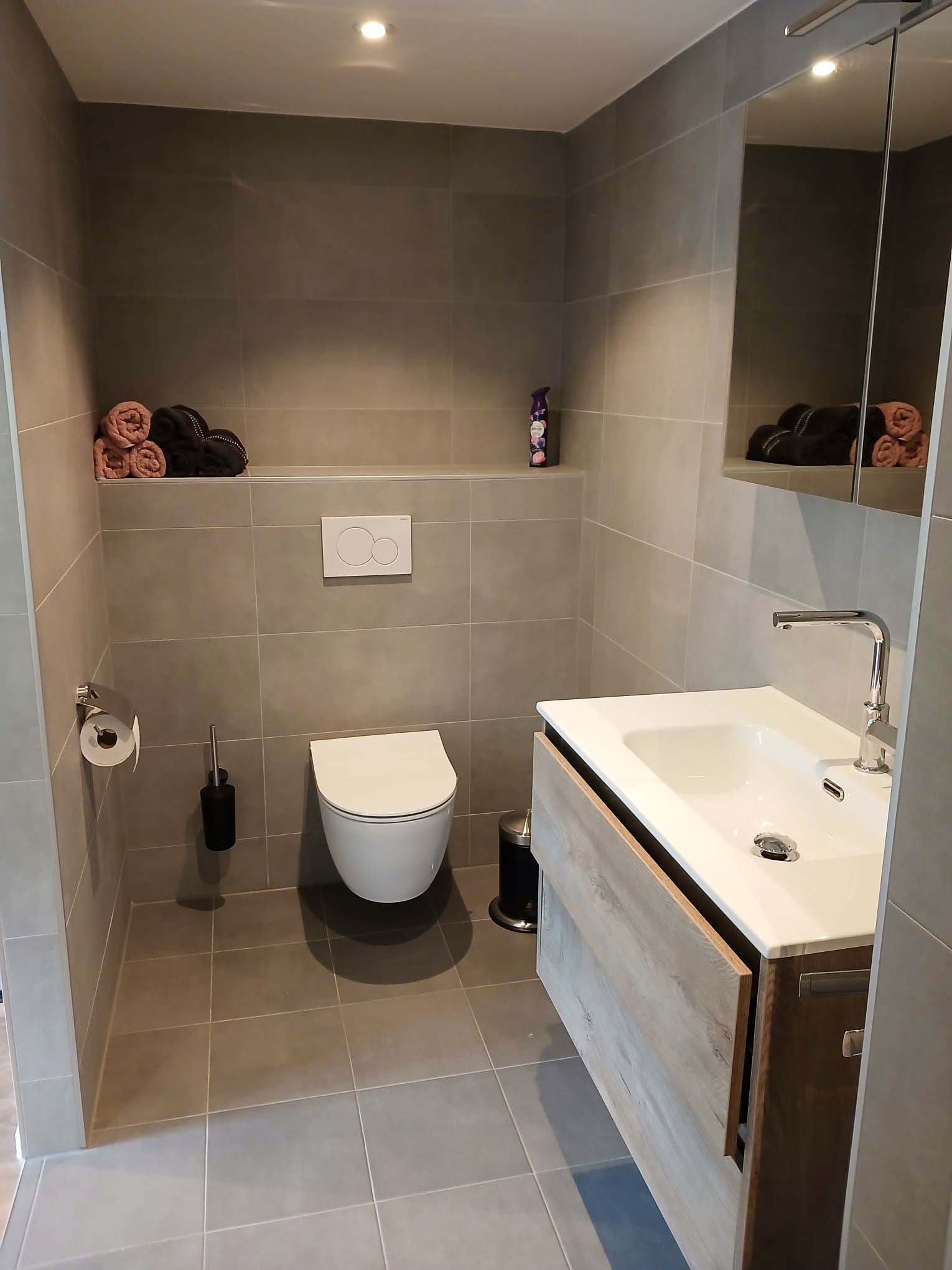 Badkamer met hangend toilet, houten wastafelmeubel en grote grijze wandtegels