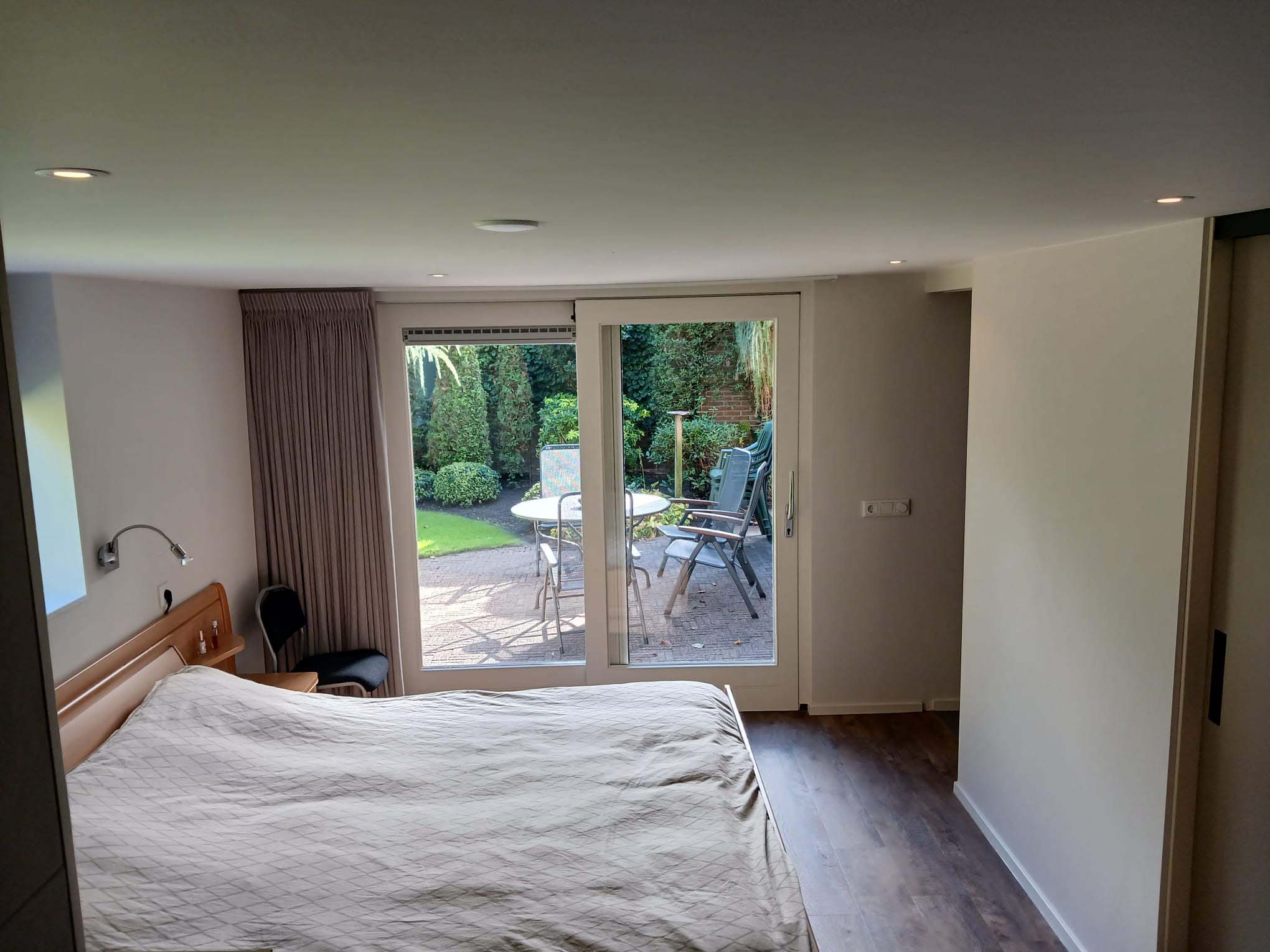 Slaapkamer na verbouwing met tweepersoonsbed, houten vloer en grote glazen schuifpui naar de tuin